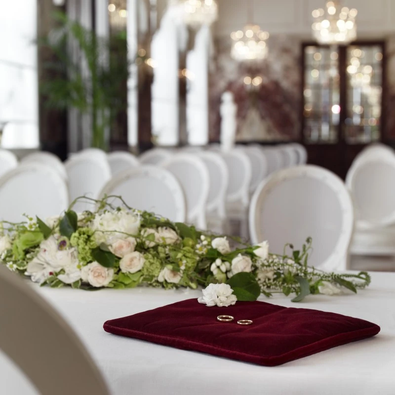 Zwei Eheringe auf einem roten Samtkissen, daneben ein elegantes Blumenarrangement aus weißen Rosen – im Hintergrund weiße Stühle und funkelnde Kronleuchter, perfekt für eine stilvolle Hochzeitszeremonie.