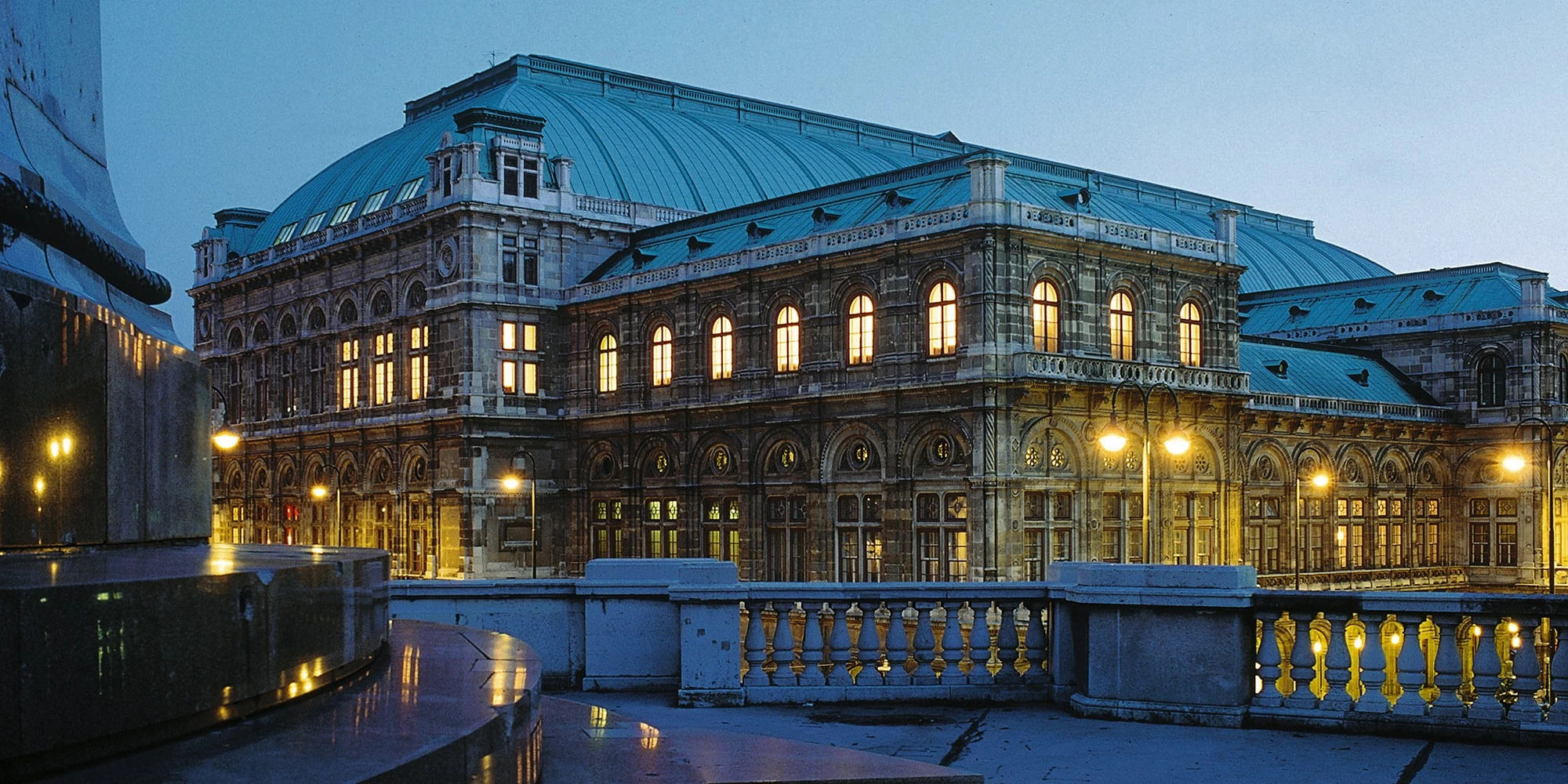Beleuchtetes Opernhaus mit markantem grünen Dach im abendlichen Licht; beeindruckende Fassade mit Rundbogenfenstern und neoromanischer Architektur.