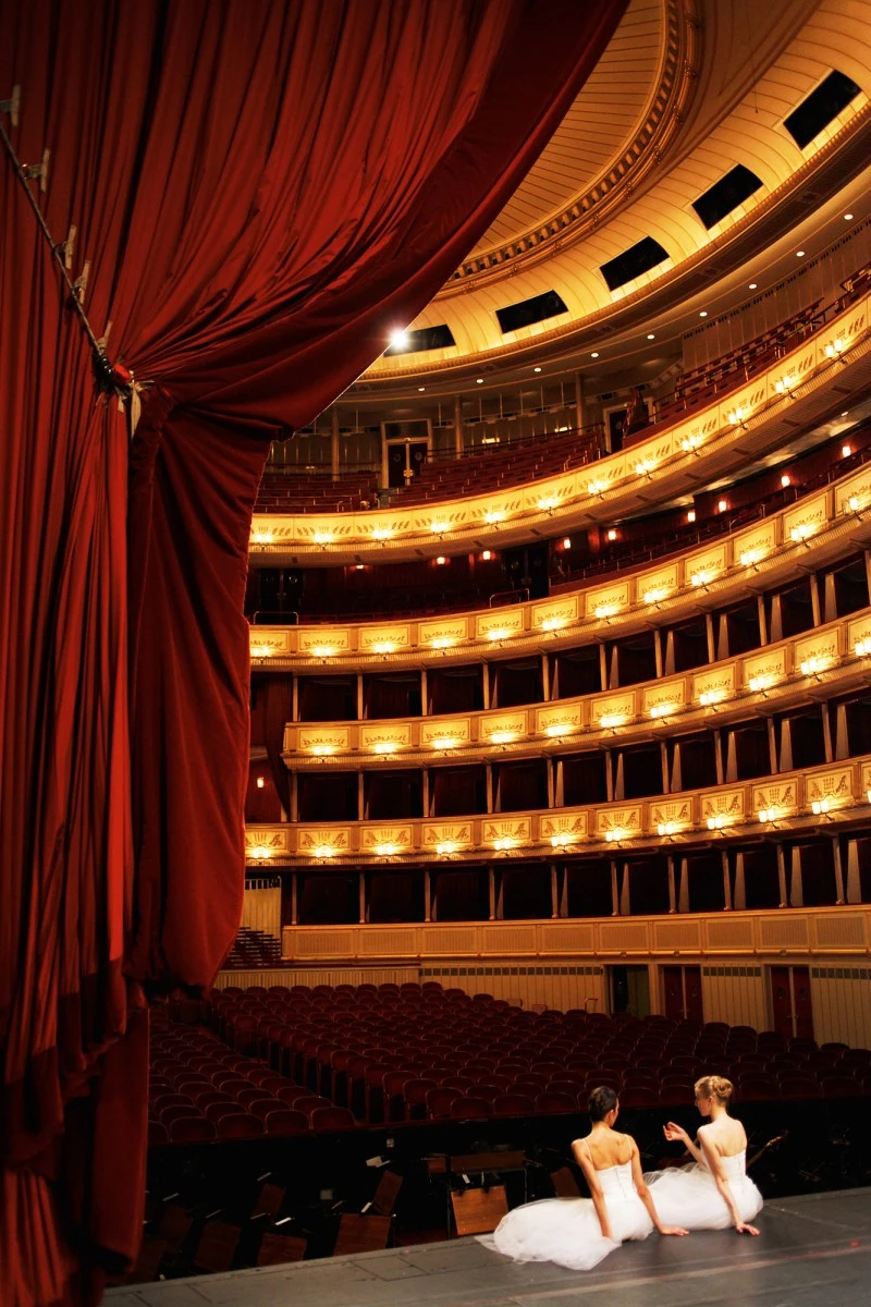 Blick von der Bühne in den prunkvollen Zuschauerraum der Wiener Staatsoper mit goldverzierten Logen, rotem Samtvorhang und zwei Tänzerinnen in weißen Tutus, die auf dem Bühnenboden sitzen.