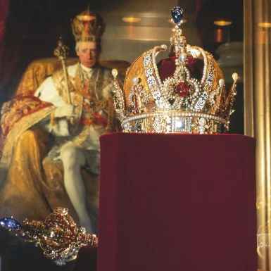 Kaiserkrone des österreichischen Kaiserreichs, reich verziert mit Gold, Edelsteinen und Perlen, im Vordergrund vor einem Porträt Kaiser Franz II./I. ausgestellt.