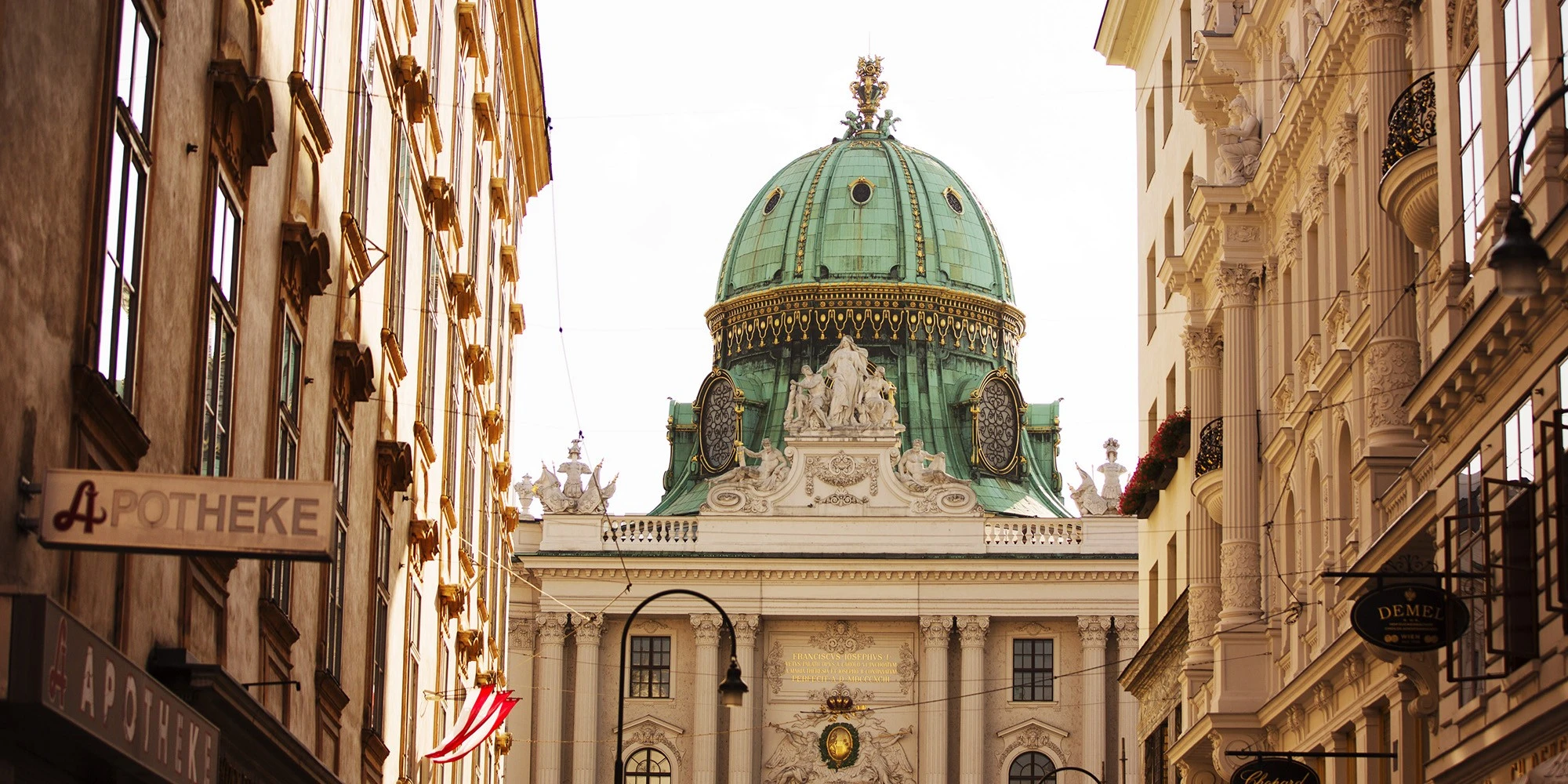 Barocke Kuppel der Hofburg in Wien, eingerahmt von historischen Altstadtfassaden mit klassischem Stuckdekor und österreichischer Flagge.