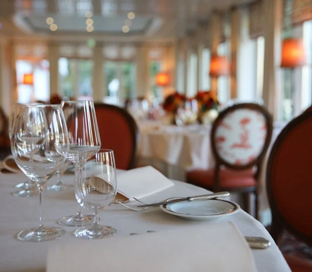 Nahaufnahme eines elegant gedeckten Tisches in einem stilvollen Restaurant mit weißer Tischdecke, feinem Glas- und Silberbesteck, im Hintergrund warme Beleuchtung und klassisch gepolsterte Stühle mit rotem Dekor.