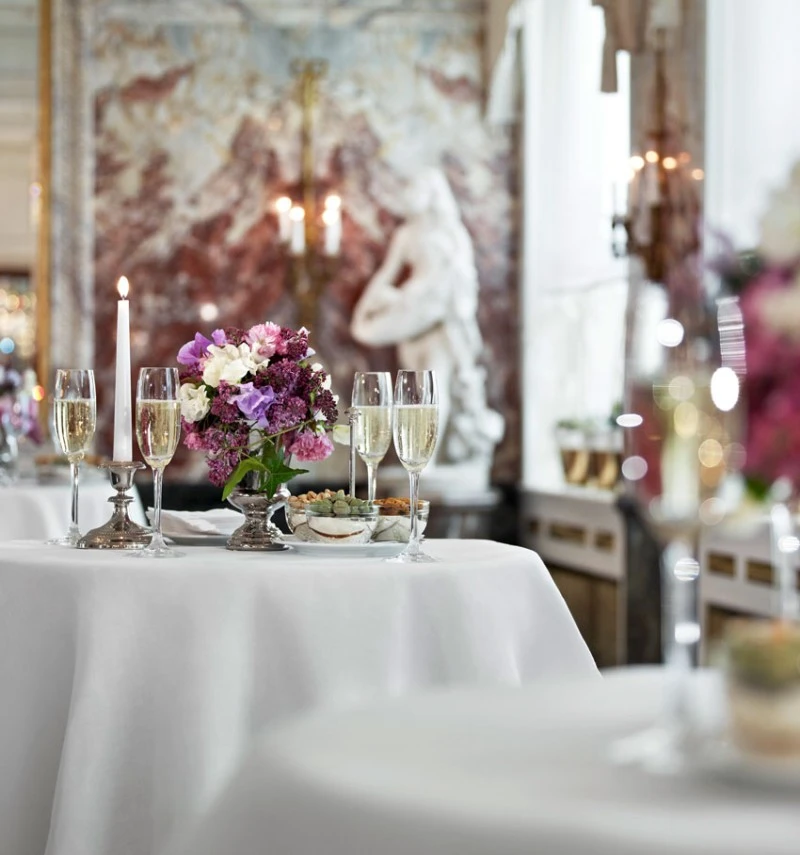Eleganter Stehtisch mit weißer Tischdecke, dekoriert mit Blumenarrangement, brennender Kerze, Champagnergläsern und kleinen Snacks, im stilvollen Marmorsaal-Ambiente.