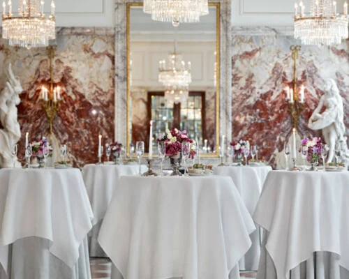 Eleganter Saal mit Marmorsäulen, großen Spiegeln, opulenten Kronleuchtern und Stehtischen mit weißen Tischdecken und floralen Arrangements.