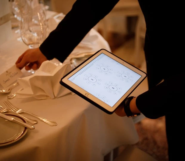 Person richtet eine festlich gedeckte Tafel ein und nutzt dabei ein Tablet mit digitalem Sitzplan zur Platzierung der Gäste.