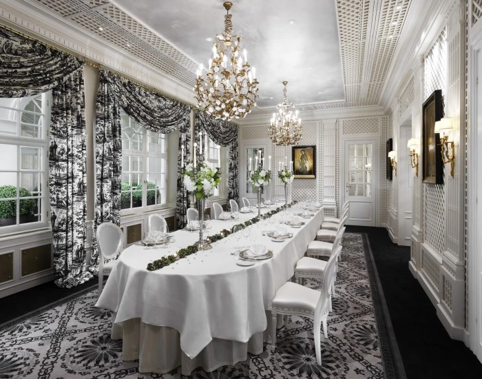Elegantes Speisezimmer mit langem, festlich gedecktem Tisch, weißen Stühlen im Louis-Seize-Stil, opulenten Kronleuchtern und schwarz-weißen Toile-de-Jouy-Vorhängen.