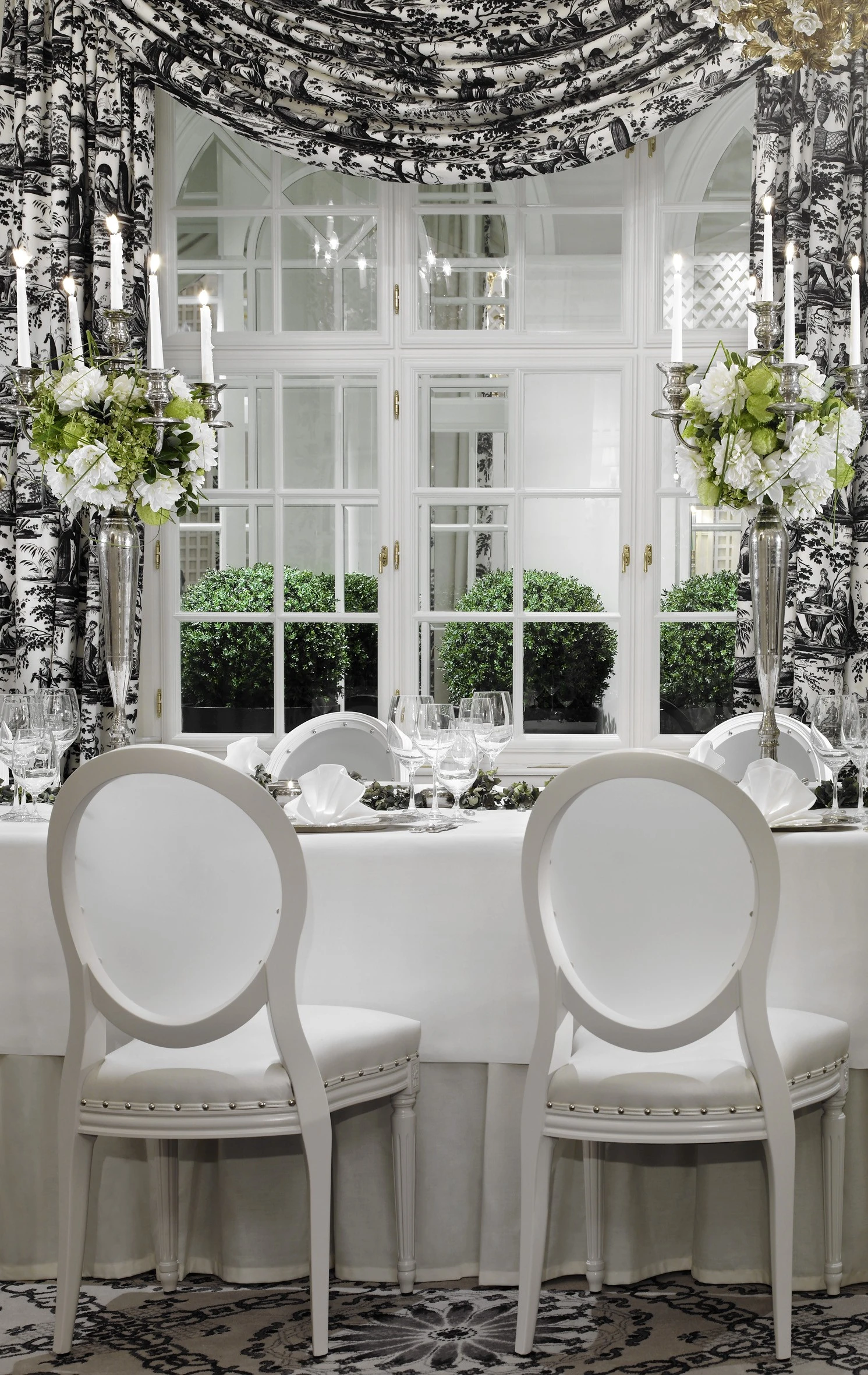 Eleganter Detailausschnitt eines klassischen Speisesaals mit weiß gedecktem Tisch, opulenten Leuchtern, silbernen Kerzenhaltern und floralen Akzenten vor schwarz-weißen Toile-de-Jouy-Vorhängen.