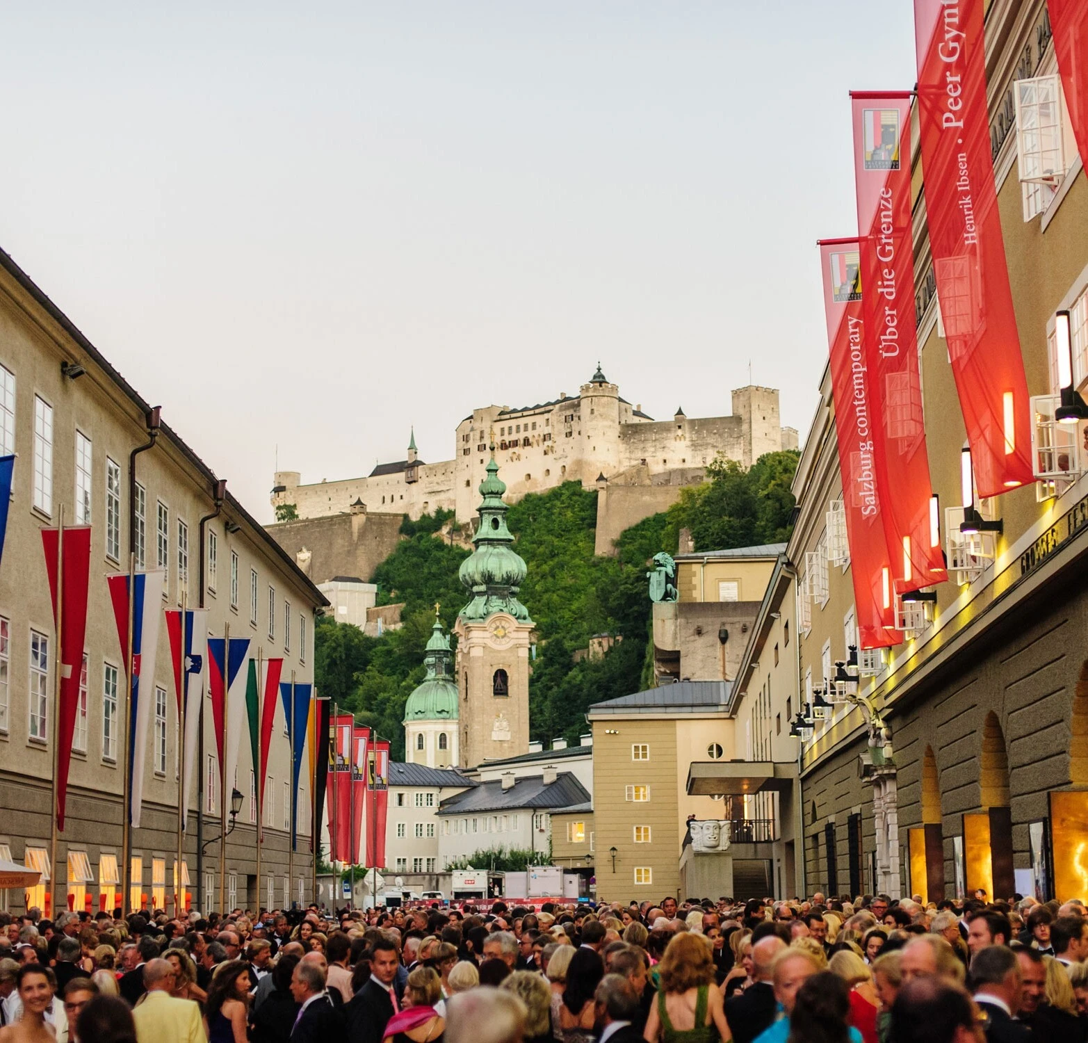 Blick auf die Festung Hohensalzburg, die imposant über den Dächern der Altstadt von Salzburg thront. Im Vordergrund barocke Kirchtürme und bunte Festspiel-Fahnen.