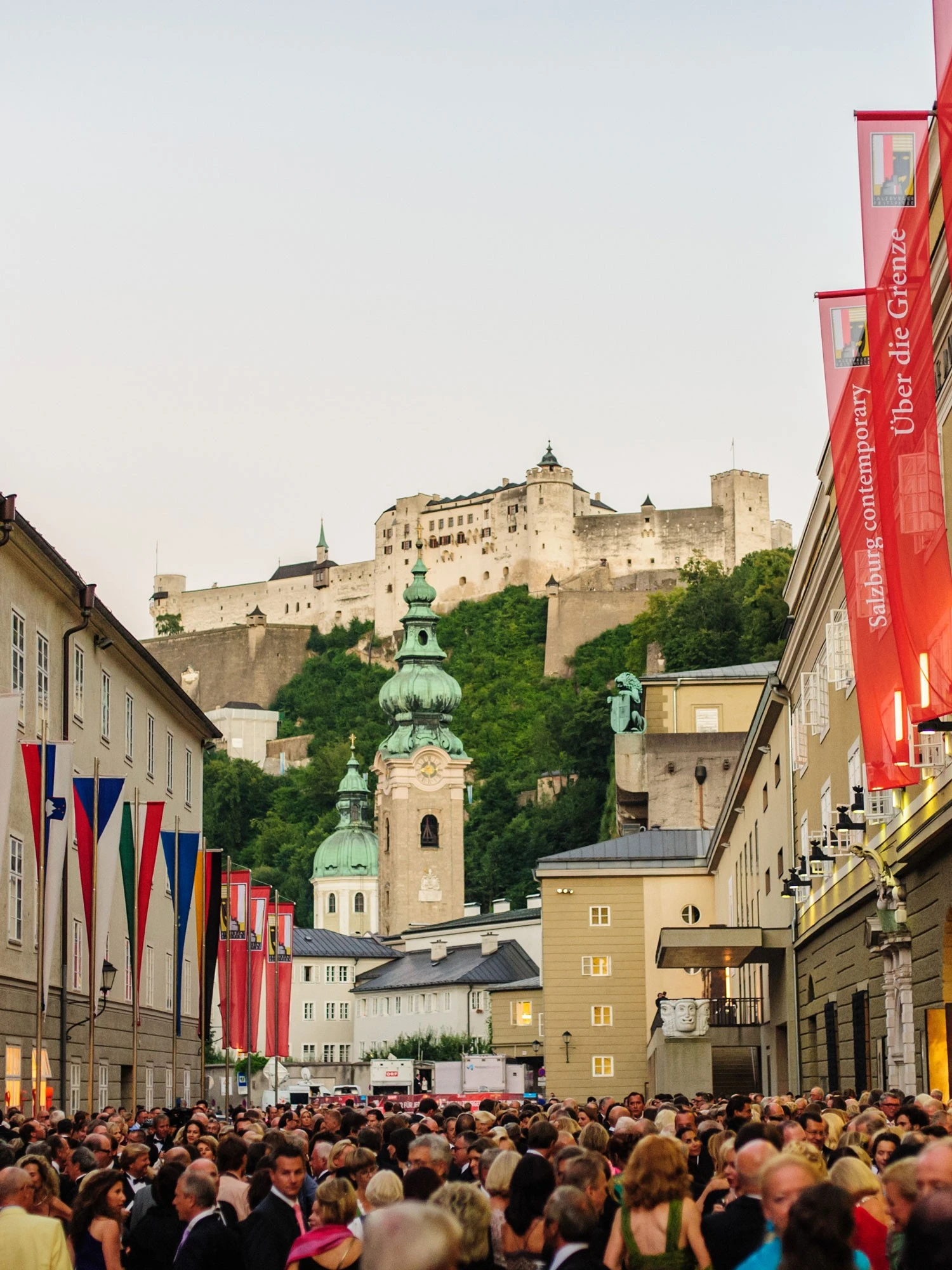 Blick auf die Festung Hohensalzburg, die imposant über den Dächern der Altstadt von Salzburg thront. Im Vordergrund barocke Kirchtürme und bunte Festspiel-Fahnen.