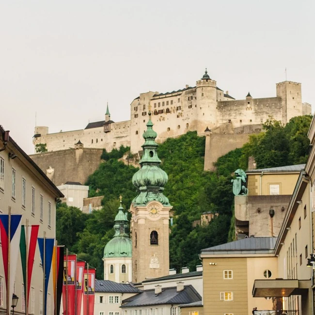 Blick auf die Festung Hohensalzburg, die imposant über den Dächern der Altstadt von Salzburg thront. Im Vordergrund barocke Kirchtürme und bunte Festspiel-Fahnen.