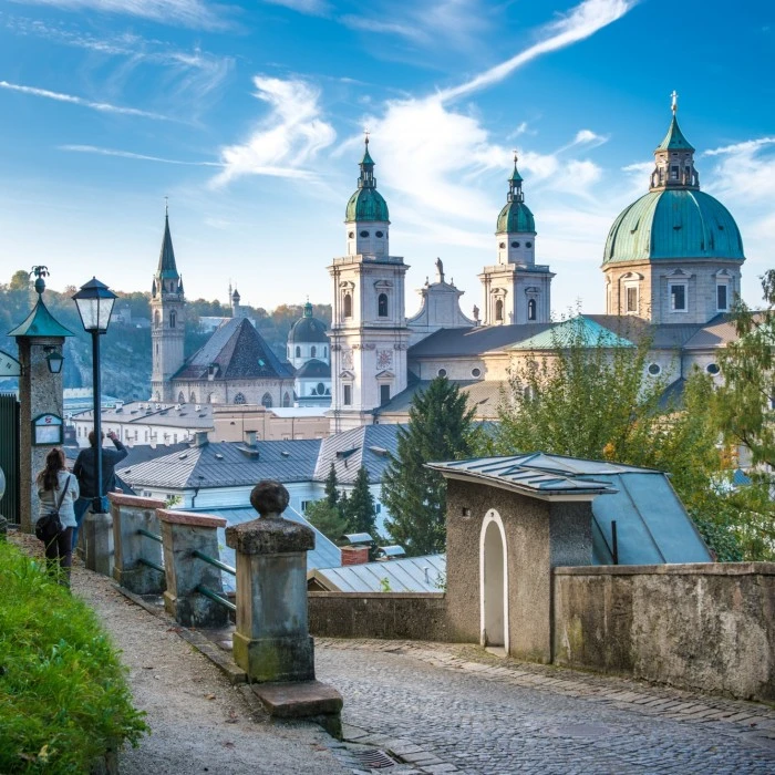 Blick von einem romantischen Fußweg mit Gastgarten-Eingang auf die barocken Türme des Salzburger Doms und umliegende Kirchen vor herbstlicher Hügellandschaft.