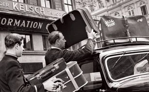 Hotel Sacher, Geschichte Zwei Männer verladen mehrere Koffer auf das Dach eines Autos vor dem historischen Eingang des Hotel Sacher. Die Szene wirkt wie eine Momentaufnahme aus den 1950er Jahren.