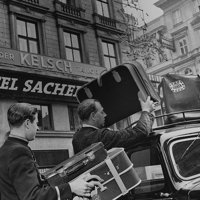 Zwei Männer laden Koffer vor dem Hotel Sacher in ein Auto. Ein Koffer trägt einen „Hotel Sacher“-Aufkleber.