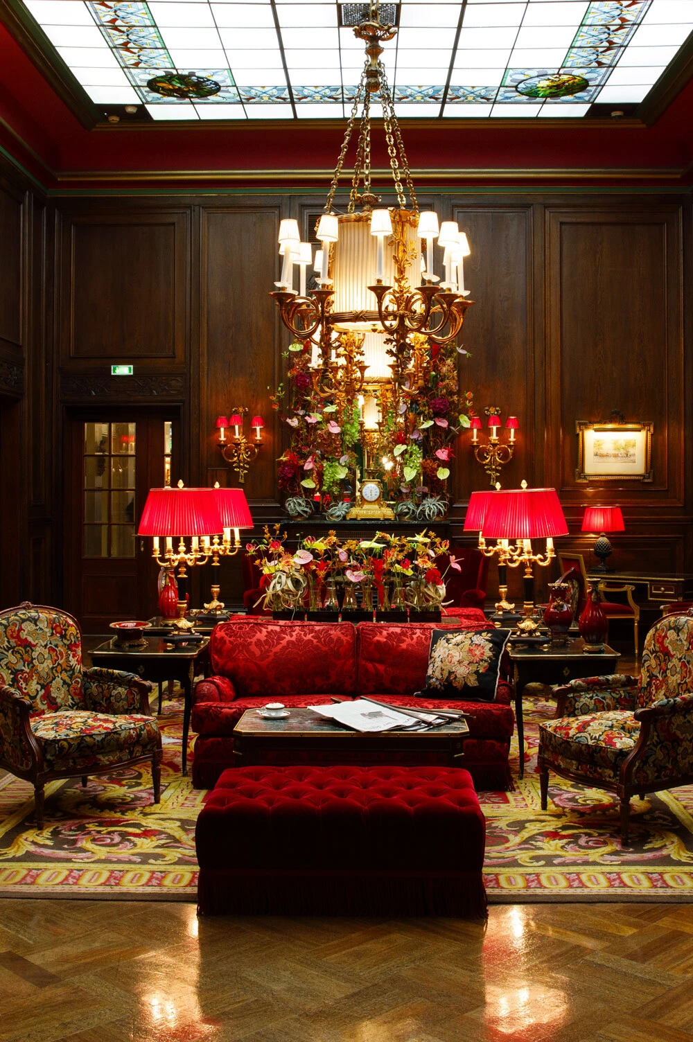 Ein prunkvoll eingerichteter Salon mit dunklem Holzvertäfelungen, rotem Samtmobiliar und floralen Mustern. Im Zentrum stehen ein großer goldener Kronleuchter, zahlreiche Blumenarrangements und elegante Leuchten mit roten Schirmen