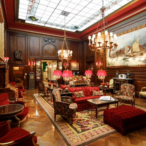 Prunkvoller Salon mit rotem Mobiliar, Parkettboden, großem Gemälde und kunstvoller Decke.