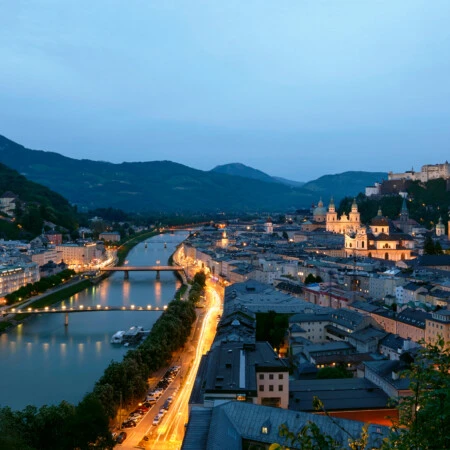 Stadtansicht von Salzburg bei Dämmerung mit der Festung Hohensalzburg, der Altstadt, dem Fluss Salzach und umliegenden Bergen.