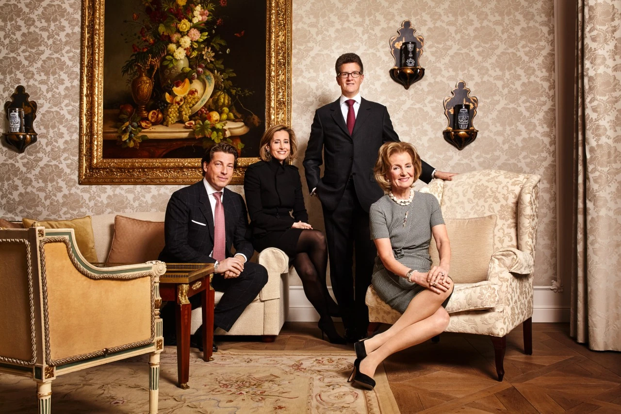 Vier elegant gekleidete Personen posieren in einem klassisch eingerichteten Raum mit gemusterten Tapeten, antiken Möbeln und einem prunkvollen Blumenstillleben in Goldrahmung. Die Szene vermittelt Tradition, Stil und familiäre Verbundenheit.