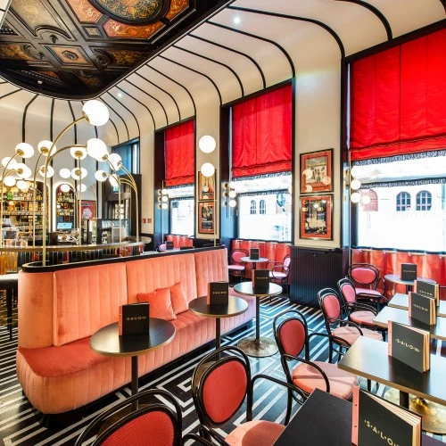 Ein stilvoll eingerichtetes Bar mit geometrischem Bodenmuster, hohen roten Vorhängen und eleganter Beleuchtung. Die Einrichtung kombiniert schwarze Polsterbänke, rosafarbene Akzente und goldene Details, ergänzt durch gerahmte Vintage-Plakate und runde Spiegel an den Wänden.