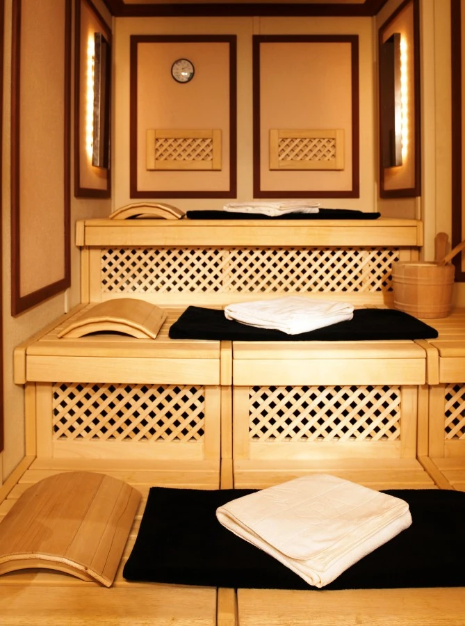 Sacher_Wien_Spa_DaySpa-Zitat-2 Mehrstufige Sauna aus hellem Holz mit schwarzen Auflagen und weißen Handtüchern, Eimer mit Schöpfkelle am Rand.