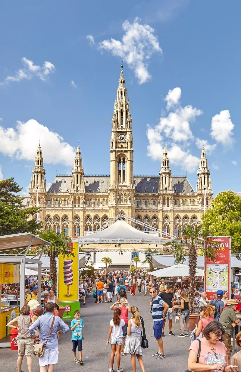 Wiener Rathaus bei Sonnenschein, davor sommerliches Fest mit bunten Ständen, Palmen, Eiswerbung und vielen Besuchern in Freizeitkleidung.