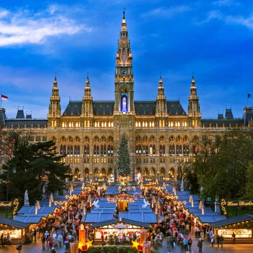 Festlich beleuchteten Wiener Christkindlmarkt vor dem eindrucksvollen Wiener Rathaus. Die Szenerie ist geprägt von festlich geschmückten Marktständen, Lichterketten und einem großen Weihnachtsbaum im Zentrum.