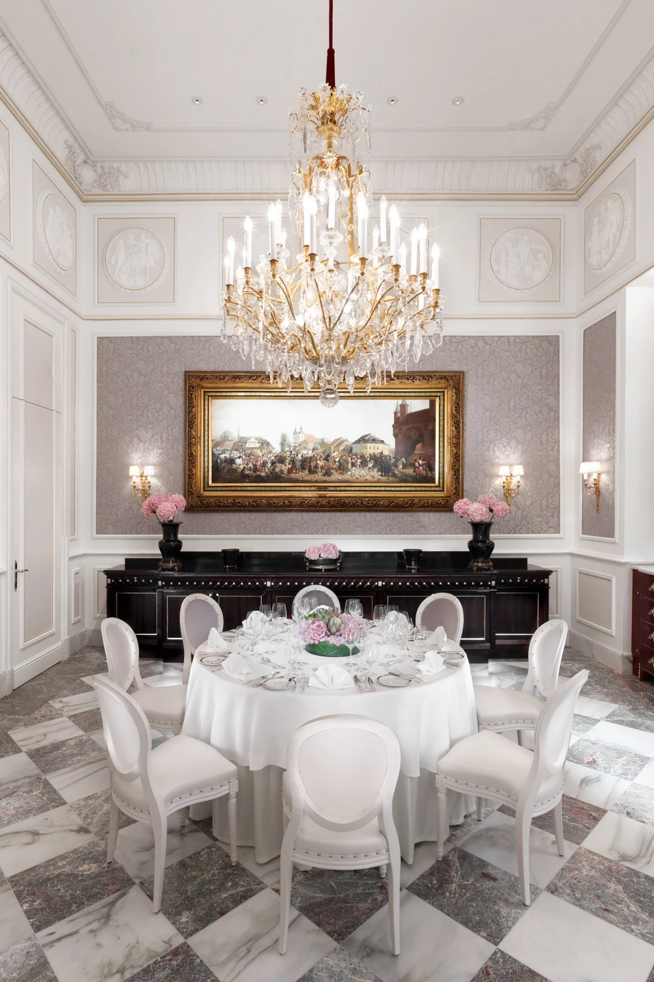 Eleganter Raum in festlichem Aufbau mit weißen Stühlen in Reihenanordnung und zentral platziertem Tisch, dekoriert mit Blumen. Wände mit lila Damasttapete, klassischer Wandverzierung und goldgerahmtem Gemälde. Ein prunkvoller Kronleuchter bildet das Zentrum der Decke.