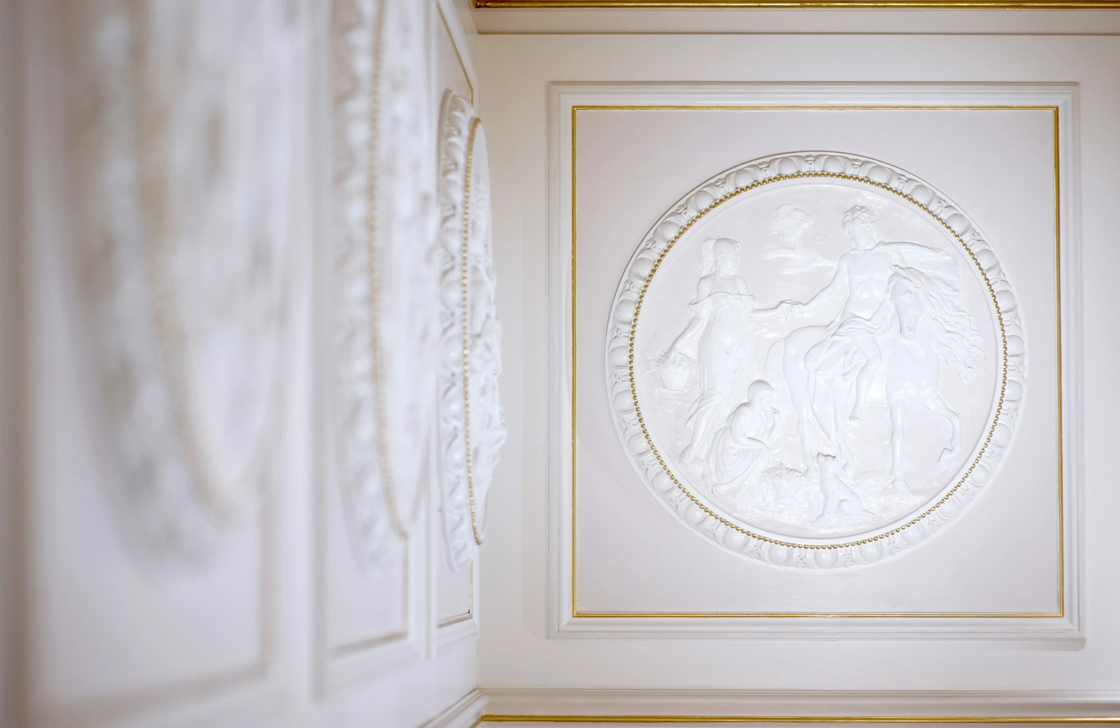 Weiße Stuckmedaillons mit fein gearbeiteten Reliefs an einer klassisch gestalteten Wand. Das zentrale Motiv zeigt eine Figur auf einem Pferd, begleitet von stehenden und knienden Personen in antikem Stil. Goldene Rahmendetails unterstreichen die Eleganz.