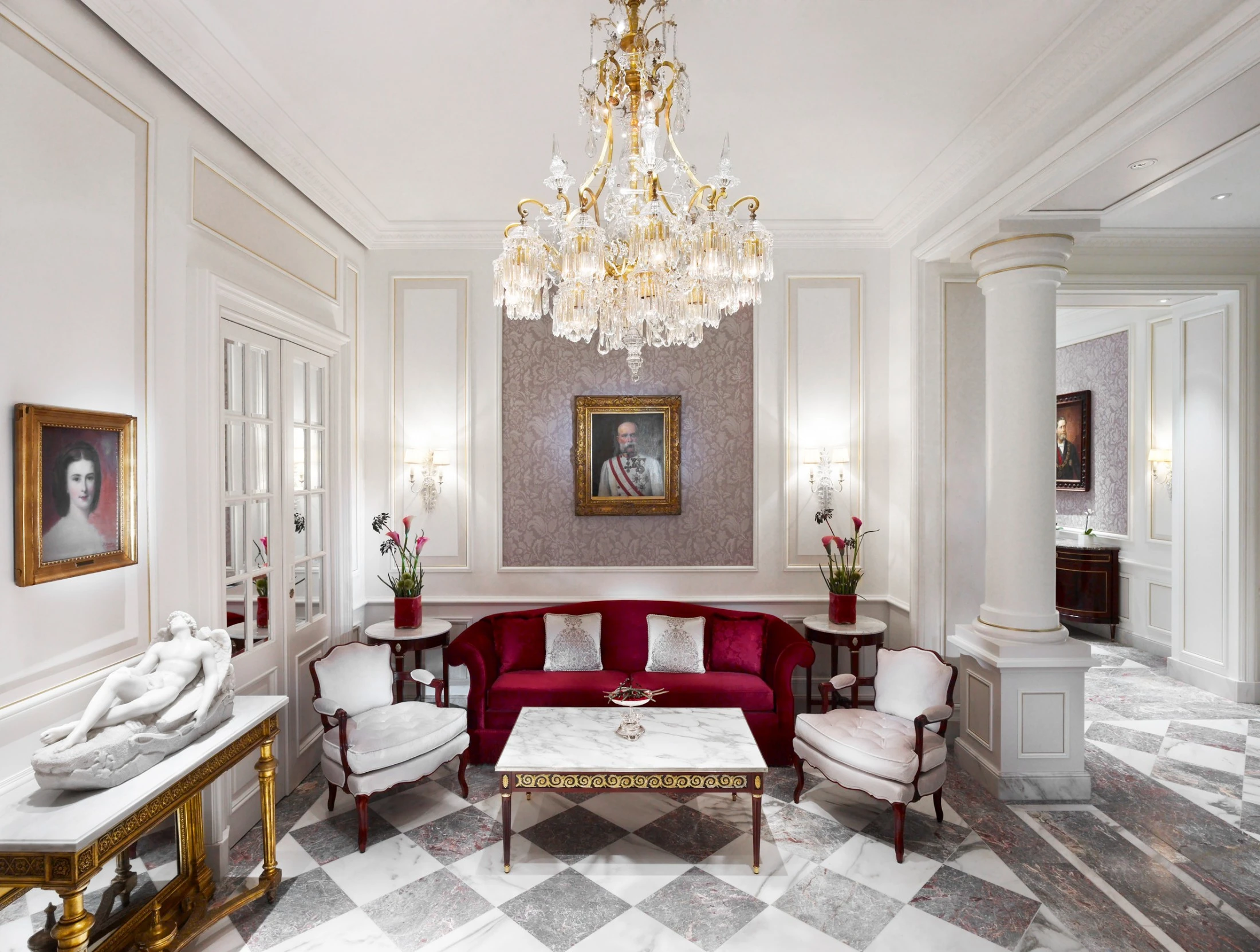 Eleganter Salon mit weiß-goldener Wandverkleidung, Marmorfußboden im Schachbrettmuster und tiefroten Samtsofas. Im Zentrum ein goldener Konsoltisch mit Marmorskulptur, darüber ein Porträt. Kristallluster, klassische Gemälde und stilvolle Möbel schaffen ein luxuriöses Ambiente.