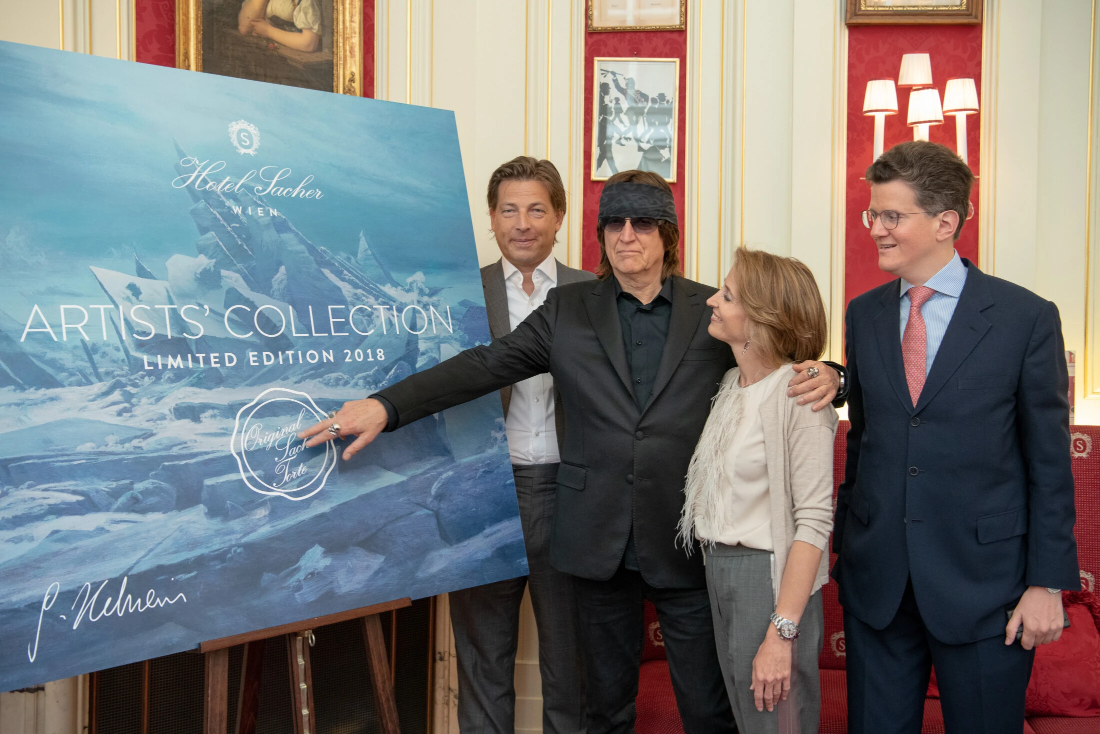 Vier elegant gekleidete Personen präsentieren ein großformatiges Plakat mit der Aufschrift „ARTISTS’ COLLECTION LIMITED EDITION 2018 – Original Sacher-Torte“. Zentrale Figur trägt Sonnenbrille und dunkle Kleidung, deutet auf das Logo. Edles Interieur mit roten Wandelementen und vergoldeten Akzenten.