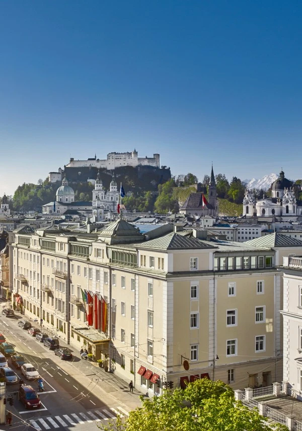 Stadtansicht von Salzburg mit der Festung Hohensalzburg auf dem Mönchsberg im Hintergrund. Im Vordergrund historische Gebäude der Altstadt, darunter ein Hotel mit mehreren internationalen Flaggen am Eingang.