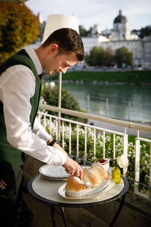 Ein Kellner in weißem Hemd, dunkelgrüner Weste und Krawatte portioniert auf einer Terrasse einen frisch gebackenen Salzburger Nockerl mit einem Löffel.