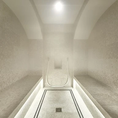 Innenansicht eines Dampfbads mit hellen, kleinformatigen Mosaikfliesen. Links und rechts befinden sich Sitzbänke, mittig zwei Duschschläuche.