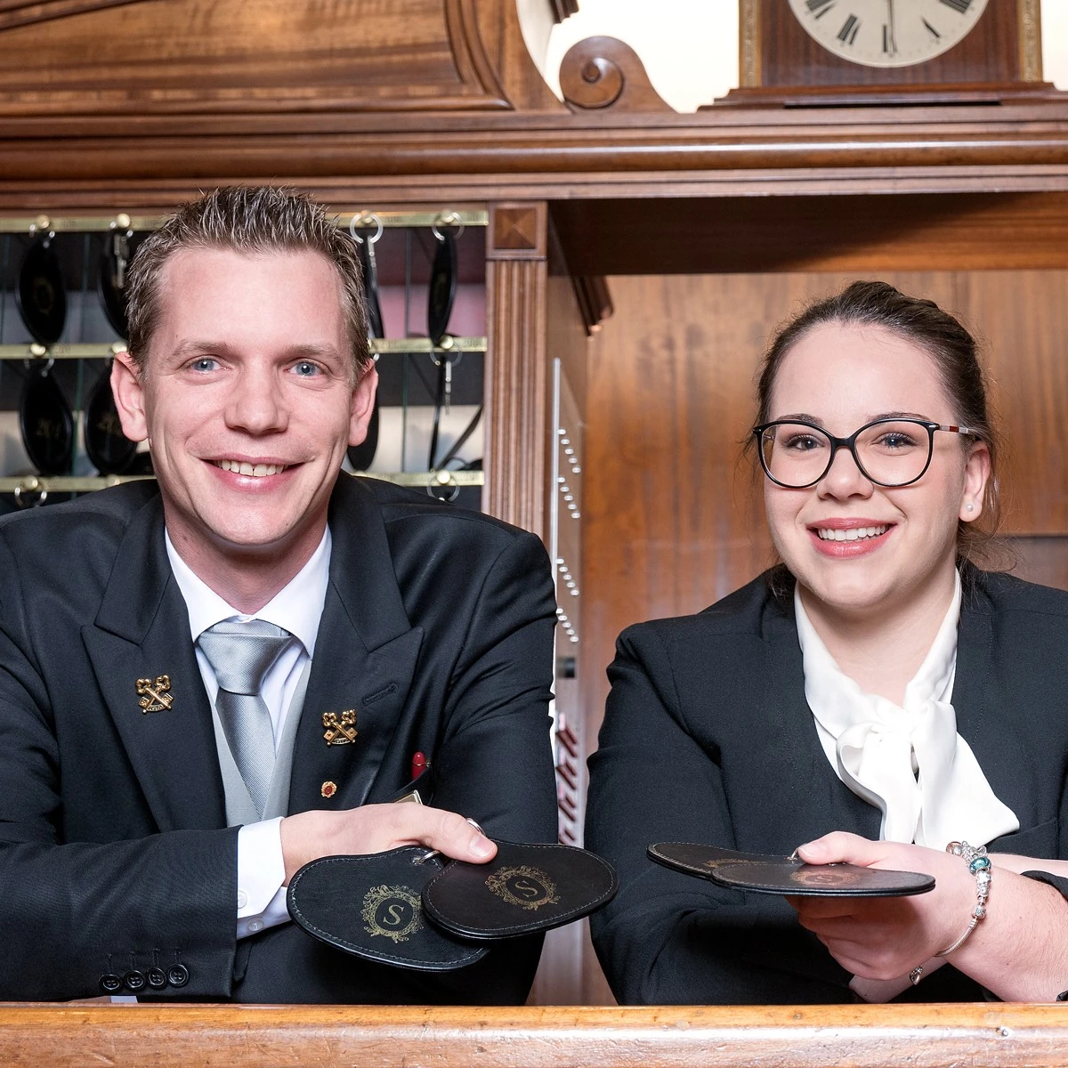 Ein Mann und eine Frau stehen hinter einem Empfangstresen, beide lächeln freundlich in die Kamera. Sie tragen elegante dunkle Kleidung mit weißen Hemden, der Mann zusätzlich eine Krawatte.
