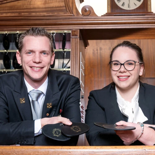 Ein Mann und eine Frau stehen hinter einem Empfangstresen, beide lächeln freundlich in die Kamera. Sie tragen elegante dunkle Kleidung mit weißen Hemden, der Mann zusätzlich eine Krawatte.