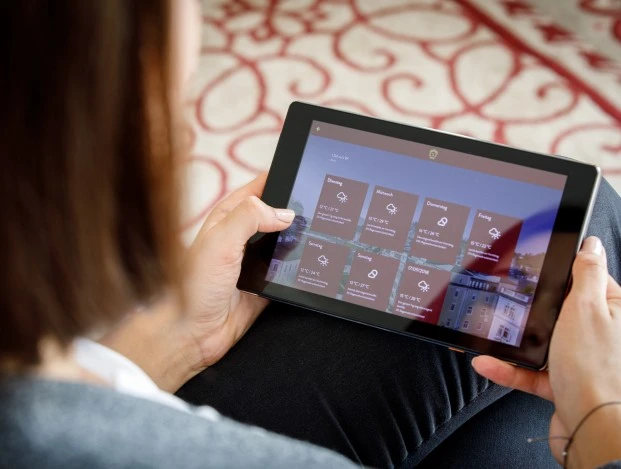 Eine Person hält ein Tablet in den Händen, auf dem eine Wetterübersicht für mehrere Tage angezeigt wird. Die Oberfläche des Tablets zeigt Temperaturen und Wetter-Symbole für verschiedene Wochentage, im Hintergrund erkennt man verschwommen eine gemusterte Oberfläche.