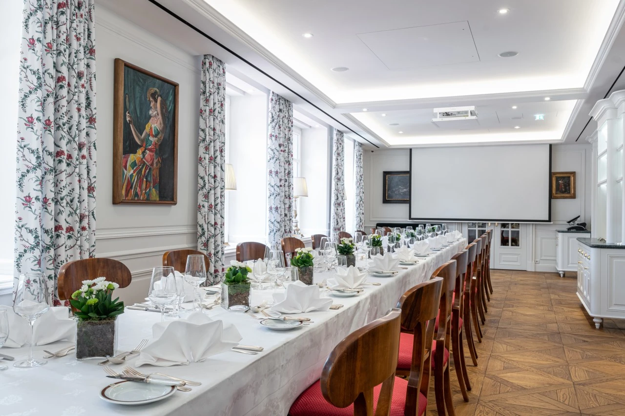 Lang gestreckter, festlich gedeckter Saal mit einer langen Tafel, weißen Stoffservietten, Porzellan, Silberbesteck, Gläsern und kleinen Blumengestecken in quadratischen Vasen.