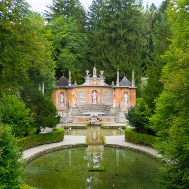 Halbrunde barocke Kulissenarchitektur inmitten eines symmetrischen Gartens mit Wasserbecken, umrahmt von dichten Bäumen. Die reich verzierte Fassade zeigt Statuen und Fresken antiker Figuren, flankiert von kleinen Türmchen und steinernen Balustraden.