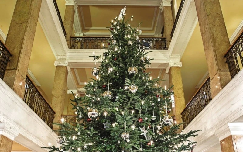 Großer, festlich geschmückter Weihnachtsbaum in einem prunkvollen Atrium mit Marmorsäulen, klassizistischen Balustraden und warmgelber Decke. Der Baum ist mit silbernen Kugeln, roten Akzenten, Sternen und funkelnden Lichtern dekoriert.