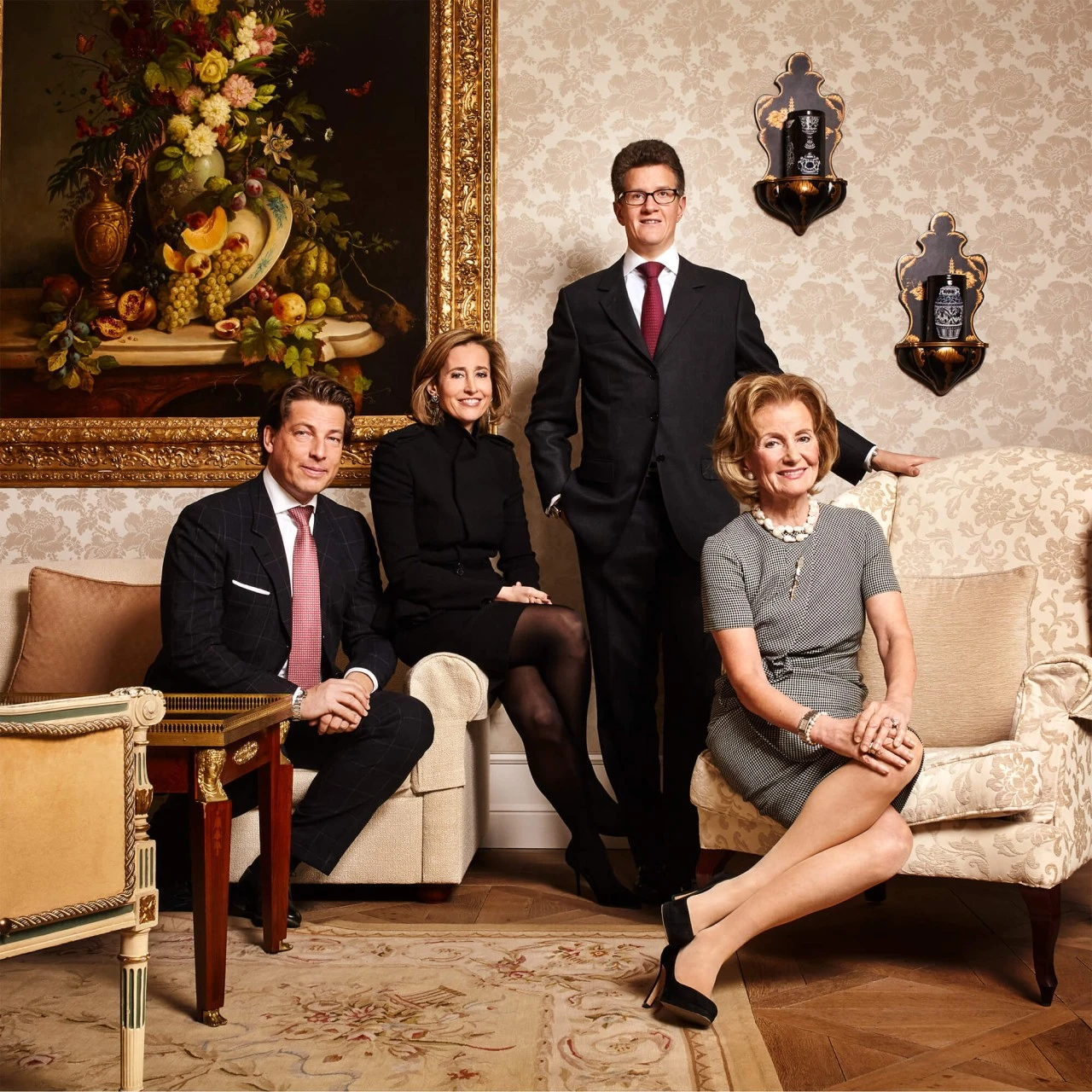 Vier elegant gekleidete Personen in klassischer Umgebung: zwei Herren im Anzug und Krawatte sowie zwei Damen, eine in Schwarz und eine in einem grauen Kleid mit Perlenkette