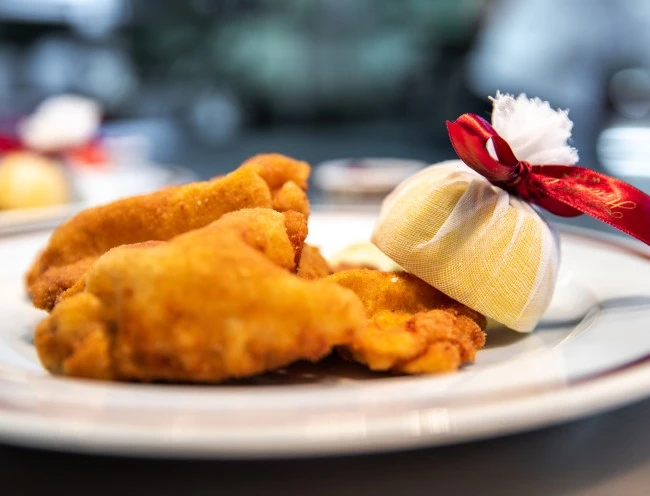 Knusprig ausgebackenes Wienerschnitzel, goldbraun paniert, mit einer in Mulltuch gewickelten Zitrone, verziert mit rotem Satinband. Serviert auf einem fein gerandeten Porzellanteller
