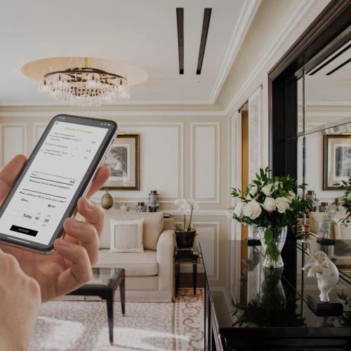 Ein Gast nutzt sein Smartphone zur Bestellung von Room Service in einer luxuriösen Hotelsuite. Auf dem Display ist die Bestellübersicht mit Speisen und Uhrzeitwahl zu sehen. Im Hintergrund eine stilvoll eingerichtete Suite mit cremefarbenen Wänden, Kristalllüster, eleganter Couch, Spiegelkonsole mit weißen Rosen und klassischem Dekor