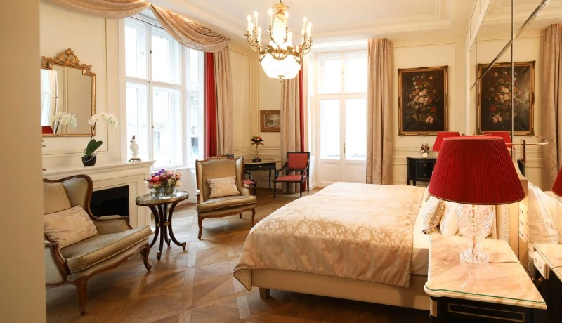 Elegantes Hotelzimmer mit klassischen Möbeln, Kristalllüster, floralen Gemälden und roten Akzenten. Helle Vorhänge und große Fenster sorgen für viel Licht.