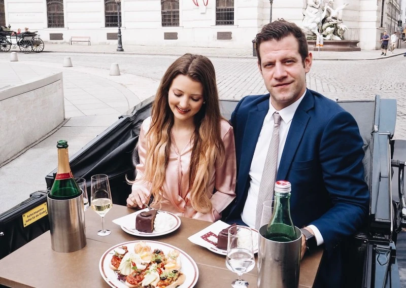 Elegant gekleidetes Paar genießt ein exklusives Essen in einer offenen Fiaker-Kutsche in der Wiener Innenstadt. Auf dem Tisch vor ihnen stehen eine Flasche Champagner, zwei Gläser, herzhafte Canapés, Sacher-Torte mit Schlagobers und Wasser.