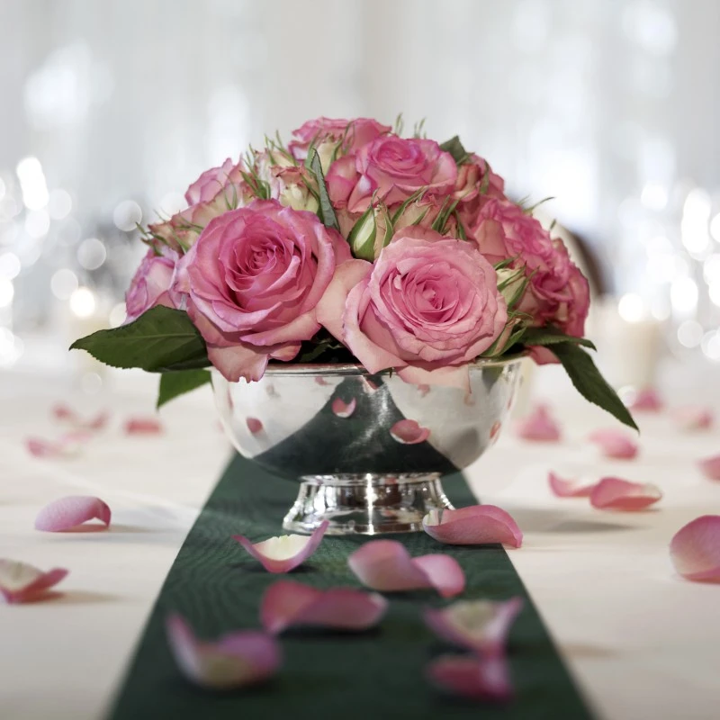 Tischdekoration mit einem kompakten Arrangement aus rosafarbenen Rosen in einer glänzenden Silbervase. Rundherum sind einzelne Rosenblätter auf einer weiß gedeckten Tafel mit grünem Mittelstreifen verstreut, im Hintergrund schimmern unscharfe Lichter und Glasreflexionen.
