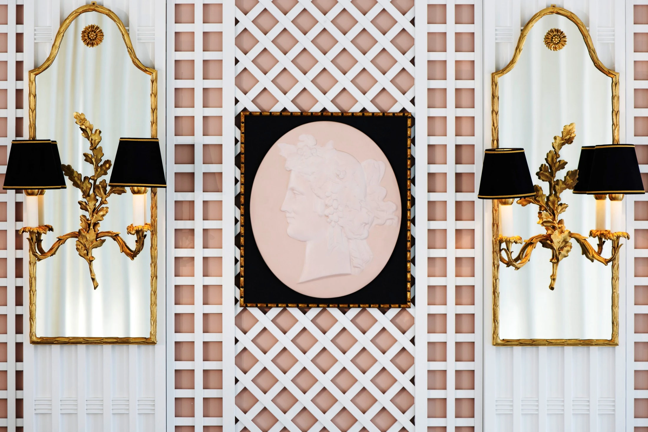 Symmetrisches Wanddekor mit zwei goldgerahmten Spiegeln, jeweils flankiert von kunstvoll vergoldeten Wandlampen mit schwarzen Schirmen. In der Mitte ein klassisches Relief eines Frauenprofils auf hellrosa Grund, gerahmt in Schwarz und Gold.