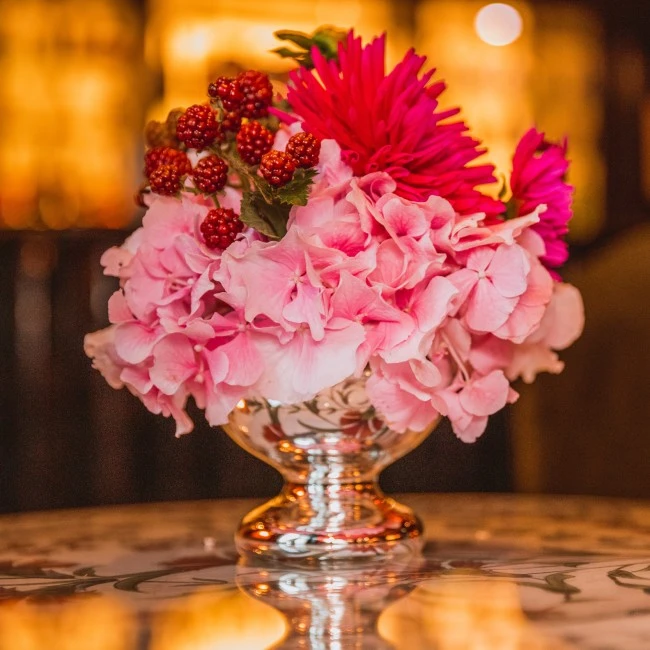 Kompaktes Blumenarrangement in einem glänzenden Silberkelch, bestehend aus zarten rosa Hortensien, kräftig pinken Blüten und dekorativen Brombeeren. Warme Hintergrundbeleuchtung sorgt für ein elegantes, stimmungsvolles Ambiente mit edler Tischreflexion.