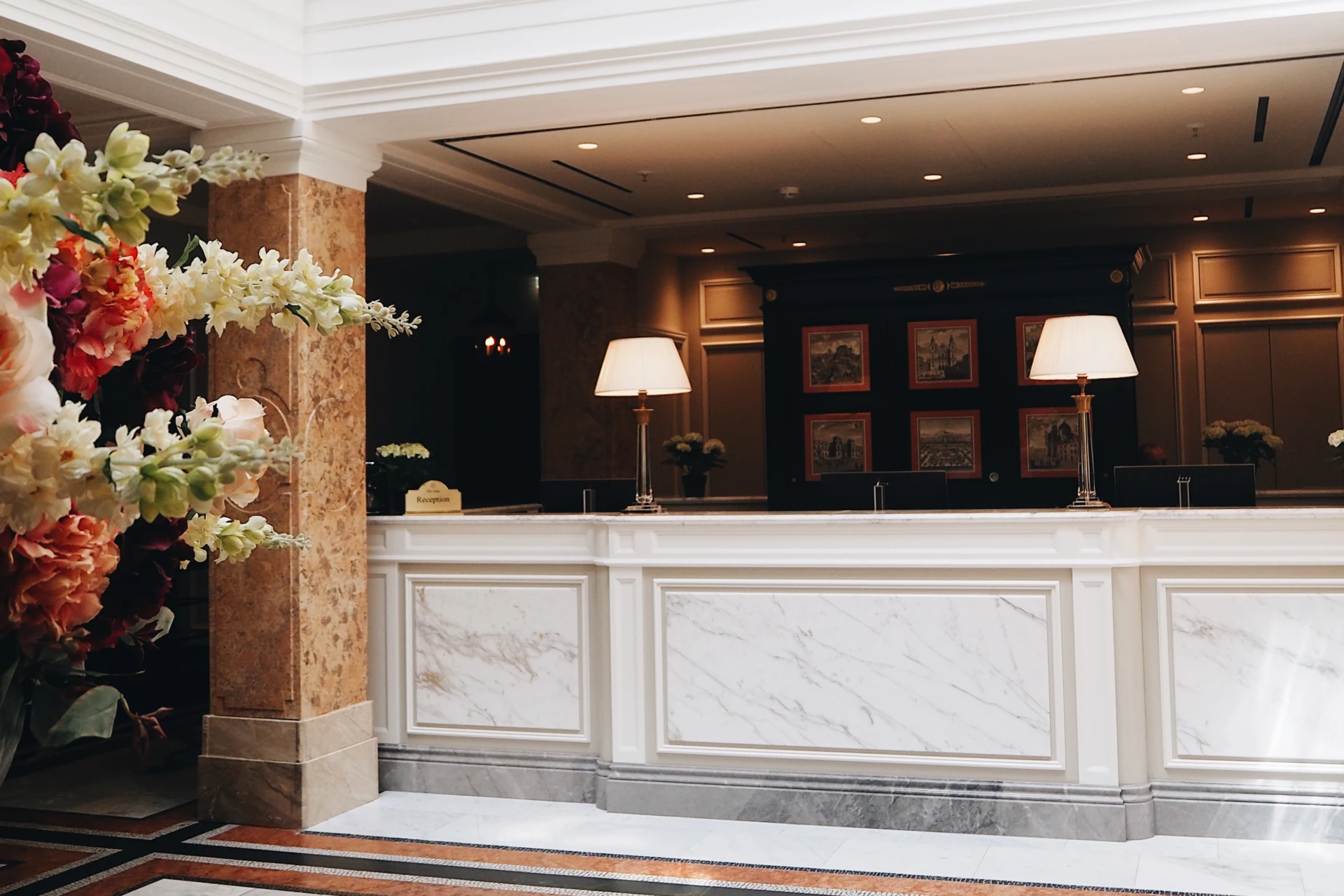 Eleganter Rezeptionsbereich mit weißem Marmor und klassischer Wandvertäfelung. Zwei stilvolle Tischlampen beleuchten die Theke, im Hintergrund gerahmte historische Stadtansichten.