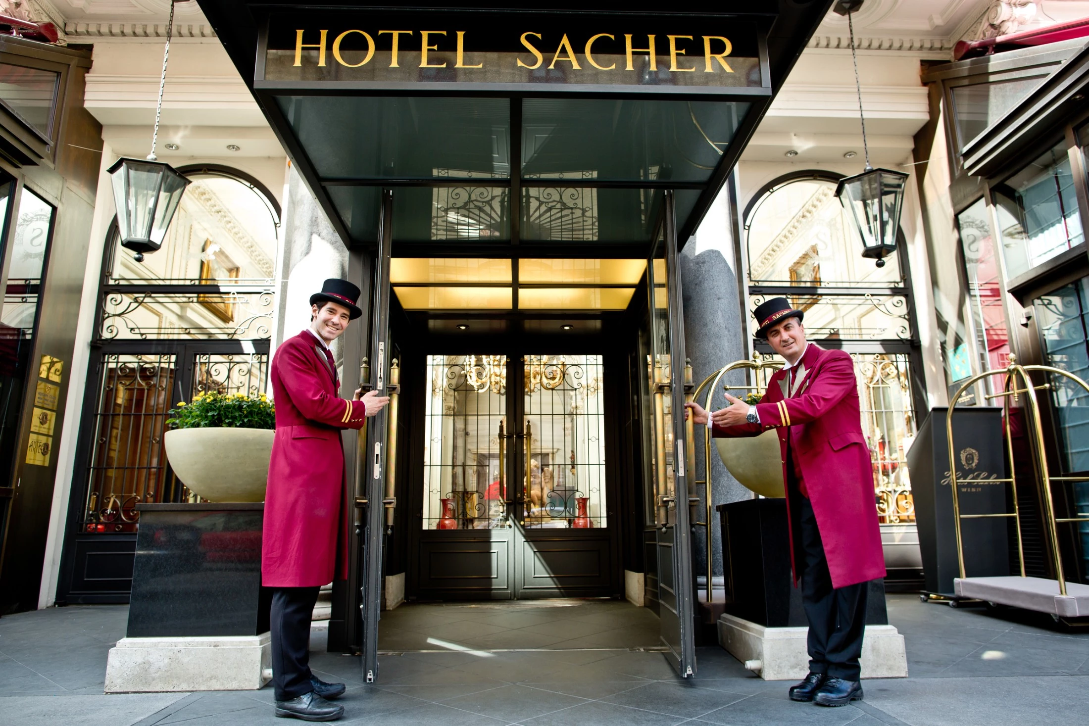 Zwei Hotelmitarbeiter in bordeauxroten Uniformen und schwarzen Hüten stehen einladend an den geöffneten Glastüren des Eingangs vom Hotel Sacher. Über ihnen prangt das goldene Schild „HOTEL SACHER“.