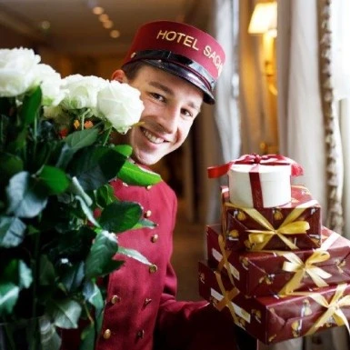 Ein lächelnder Hotelmitarbeiter in bordeauxroter Uniform mit der Aufschrift „Hotel Sacher“ auf der Mütze hält einen Strauß weißer Rosen und mehrere rot-goldene Geschenkboxen, die mit Schleifen verziert sind.