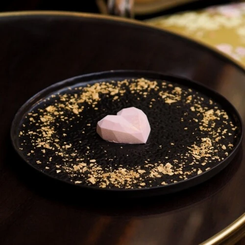 Ein rosa, geometrisch facettiertes Herz liegt mittig auf einem schwarzen Teller. Um das Herz herum sind goldene Brösel oder Splitter dekorativ verteilt. Der Teller steht auf einem dunklen Holztisch mit goldenem Rand.
